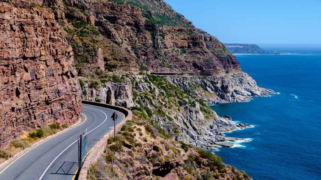 A winding coastal road curves around rocky cliffs, overlooking the blue ocean. The clear sky adds to the scenic view, and lush greenery is visible along the cliffs.