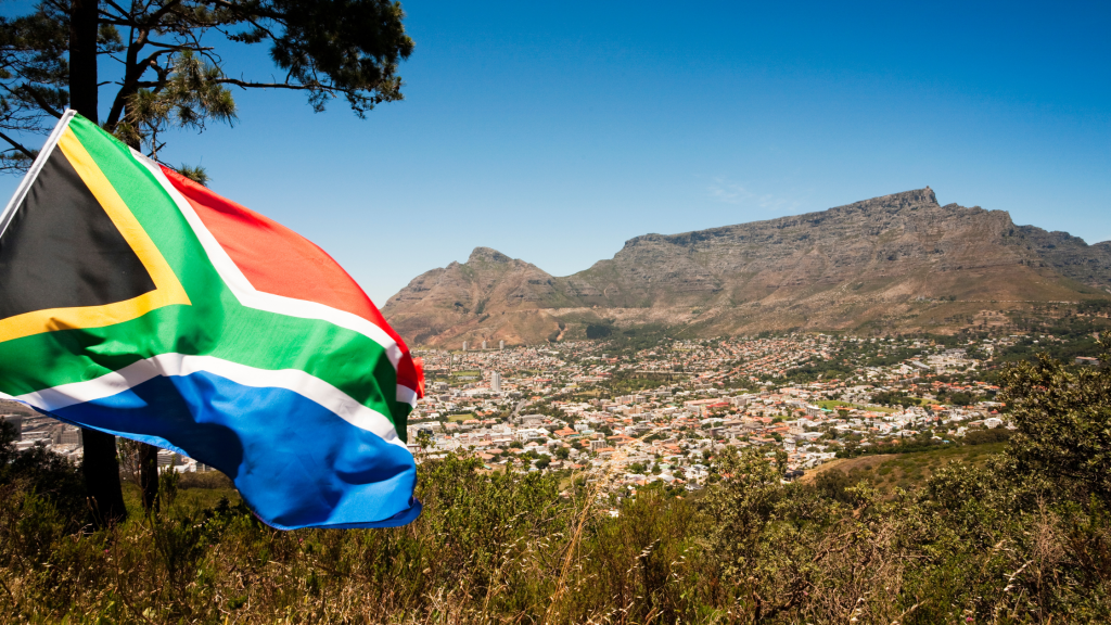 The image shows the South African flag waving in the foreground with a cityscape below. In the background, the iconic Table Mountain stands under a clear blue sky. Trees and greenery are visible around the scene.