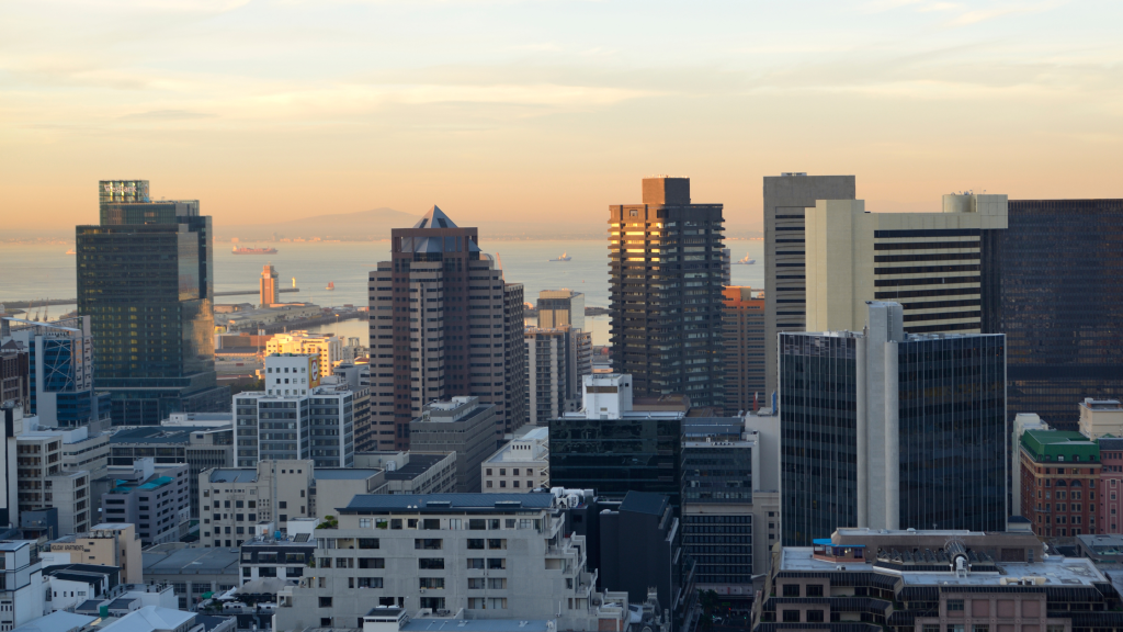 City skyline at sunset with tall buildings and a distant view of the ocean. The sky is painted with warm hues, and several ships are visible on the water. The scene captures an urban landscape transitioning into evening.