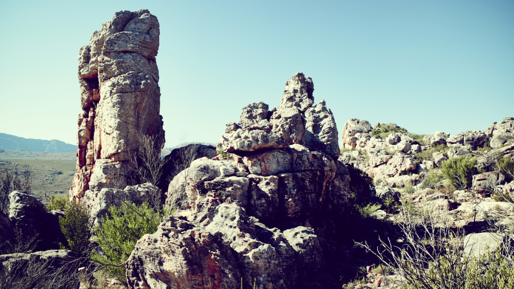 A rugged landscape featuring tall, weathered rock formations under a clear blue sky. Sparse vegetation grows among the rocks, and distant mountains are visible in the background.