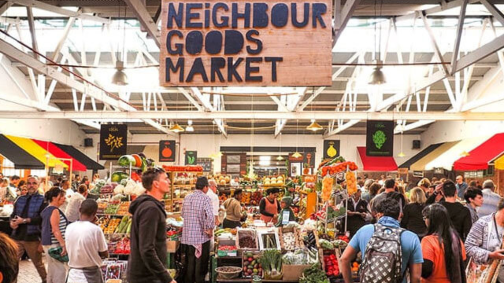 A bustling indoor market scene with people browsing various colorful stalls. A large wooden sign above reads Neighbour Goods Market. Bright banners and fresh produce are visible, creating a lively and vibrant atmosphere.