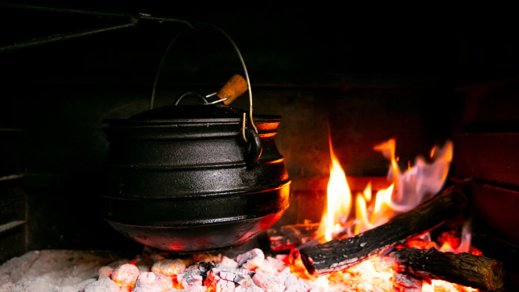 A black cast iron pot hangs over a fire in a fireplace, surrounded by glowing embers and burning logs. The flames cast a warm light on the scene, creating a rustic and cozy atmosphere.