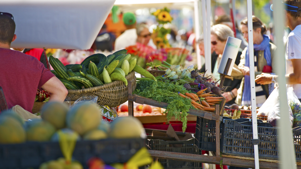 A lively farmers market scene with baskets of fresh cucumbers, carrots, and beets in the foreground. People are browsing the stalls filled with vibrant produce in the background under sunny skies.