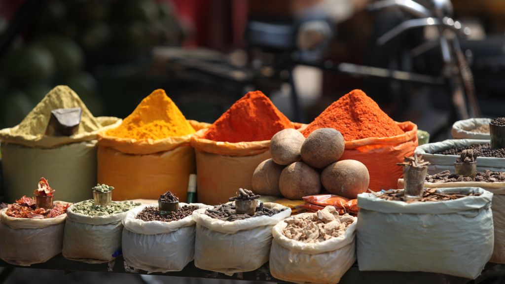 A vibrant display of spices and herbs at a market stall. Mounds of colorful powders in hues of yellow, orange, and red are surrounded by various other spices and nuts in cloth sacks, creating a lively and aromatic scene.