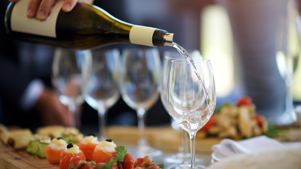 A close-up of a hand pouring white wine into a glass on a wooden table. Surrounding the wine glasses are an assortment of appetizers and small dishes, creating an elegant and inviting setting with soft, natural lighting.