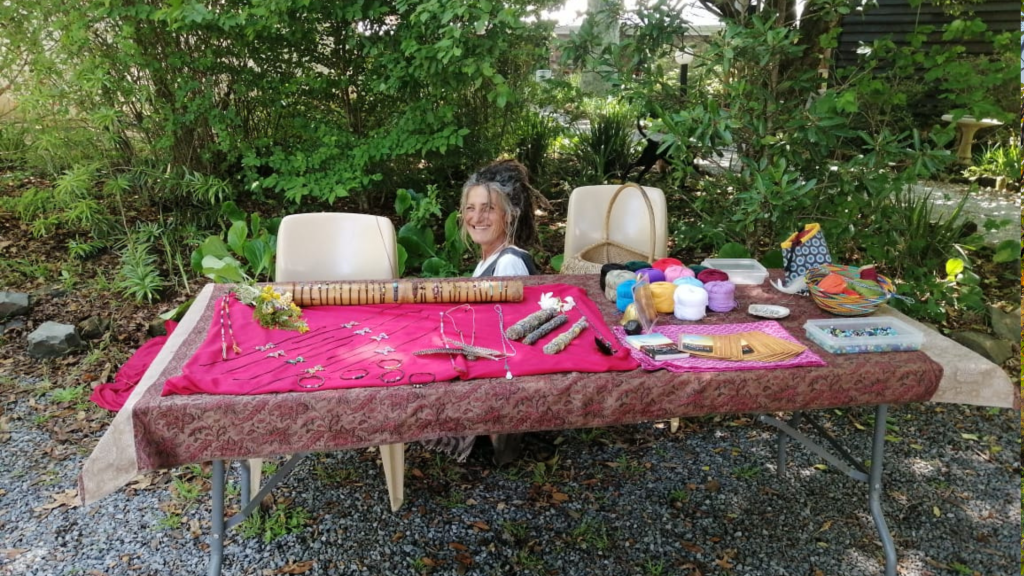 A woman seated behind a table covered with a red cloth, displaying various handmade crafts, yarn, and beads. The setting is outdoors with lush green foliage in the background.