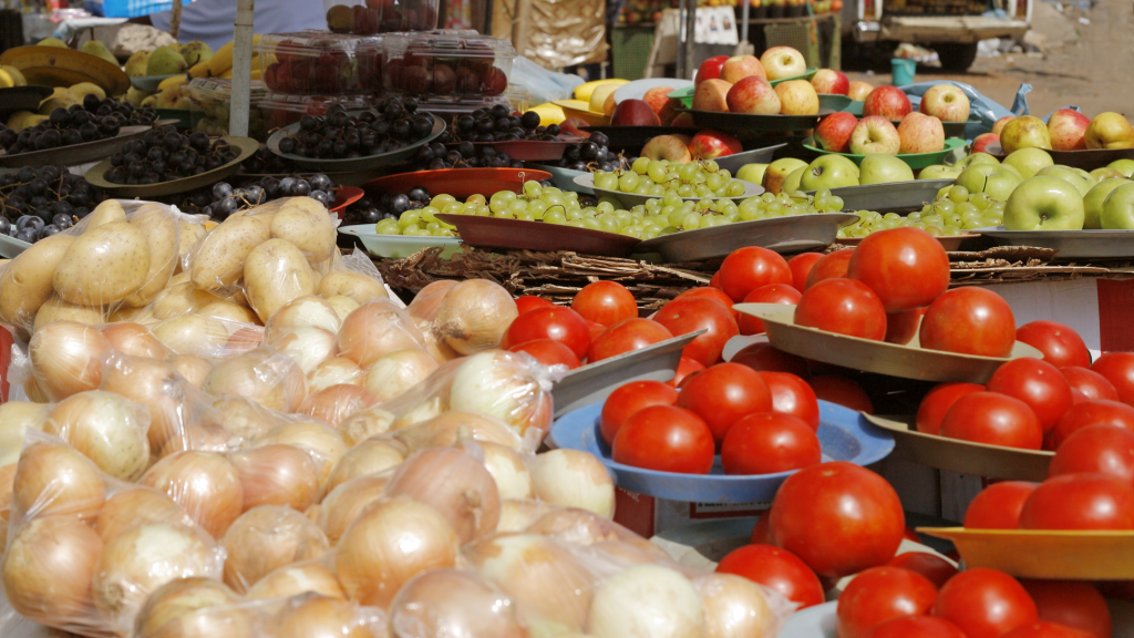 A vibrant market stall displays a variety of fresh produce, including onions, tomatoes, grapes, apples, and pears. Each type of fruit and vegetable is arranged neatly in trays, showcasing a colorful array of options for shoppers.