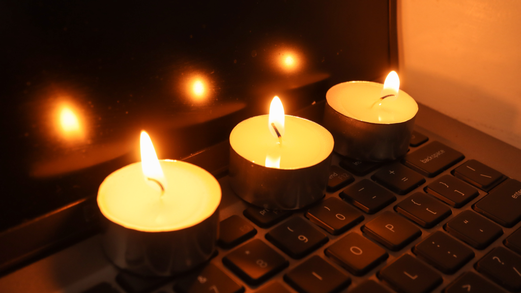 Three small candles are lit, placed on the keyboard of a laptop. The screen reflects their warm glow, creating a cozy atmosphere. The keys are partially visible, emphasizing the contrast between technology and candlelight.