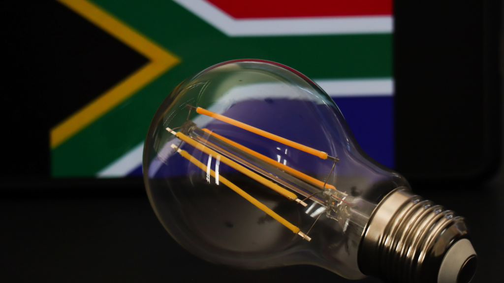 A clear LED light bulb placed in front of a blurred South African flag. The bulbs filaments are visible, symbolizing innovation and energy against a national backdrop.