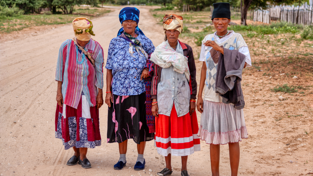 Four elderly women stand on a dirt road in colorful, traditional clothing and headscarves. They are outdoors with trees and a fence in the background. The setting appears rural and peaceful.