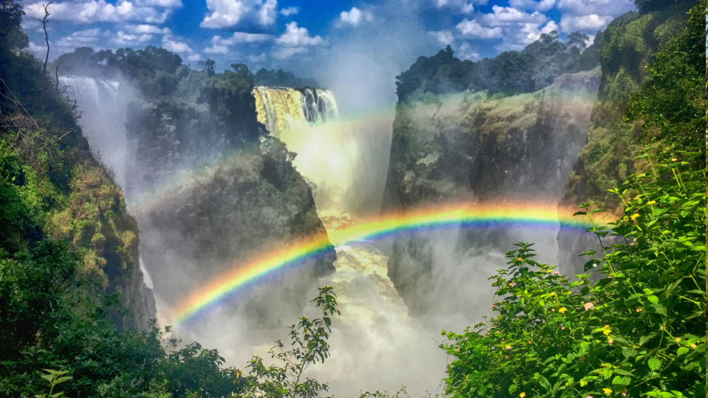 A powerful waterfall surrounded by lush greenery with mist rising into the air. Two vibrant rainbows arch over the chasm, with blue skies and scattered clouds above, creating a vivid and dynamic natural scene.