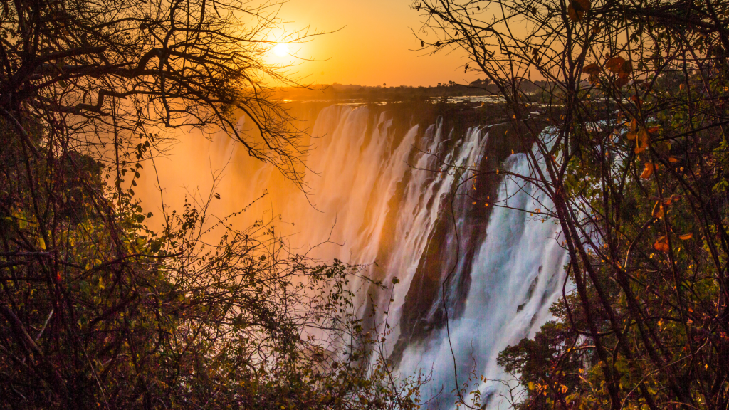 A breathtaking view of a waterfall at sunset, with cascading water illuminated by the orange glow of the sun. The scene is framed by silhouetted trees and foliage, highlighting the dramatic natural beauty of the landscape.