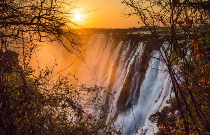 A stunning view of a waterfall cascading over a rocky cliff at sunset, surrounded by lush green foliage. The golden light creates a mist, adding a warm glow to the scene.