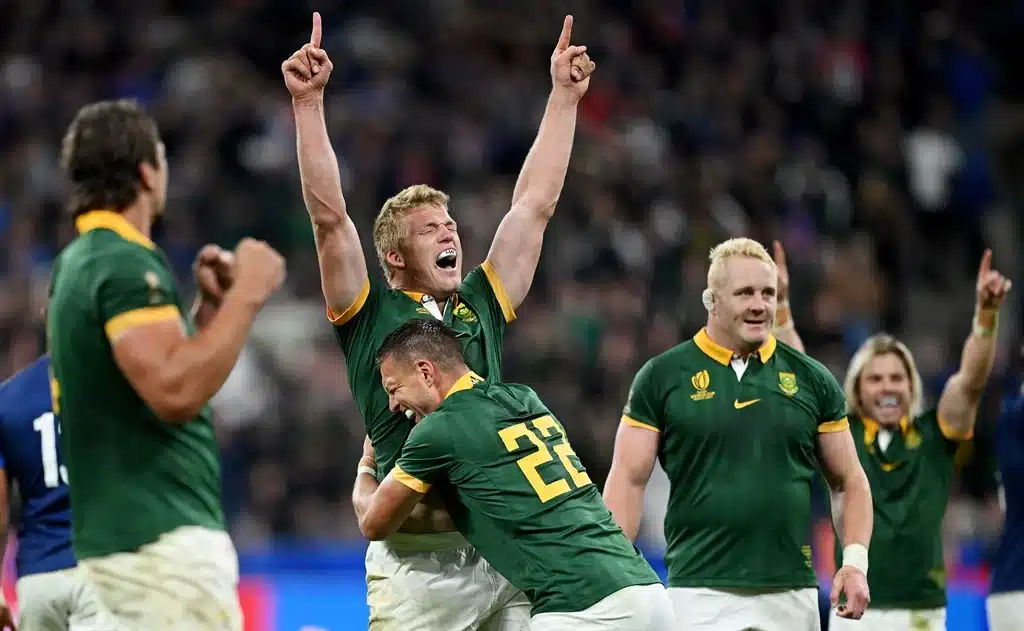 Rugby players in green jerseys celebrate a victory on the field. One player stands with his arms raised in triumph while another hugs him. Other teammates smile and clap in the background.