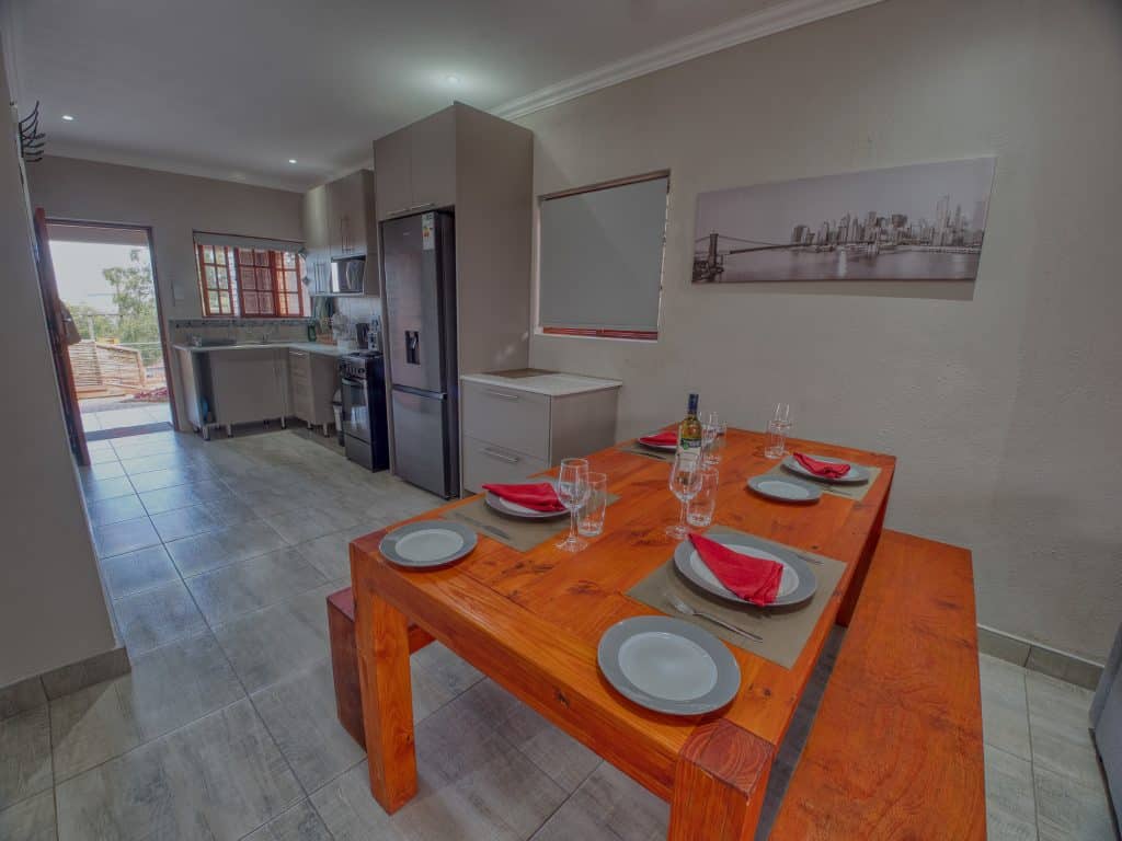 A modern kitchen and dining area featuring a wooden table set with four plates, red napkins, wine glasses, and a bottle of wine. The room includes a refrigerator, oven, and a panoramic cityscape photo on the wall, with a view to an outdoor space.