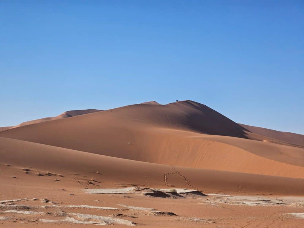 A vast desert landscape with tall, reddish sand dunes under a clear blue sky. Footprints lead across the sand towards the dunes, suggesting human activity. Sparse patches of dried, cracked earth are visible in the foreground.