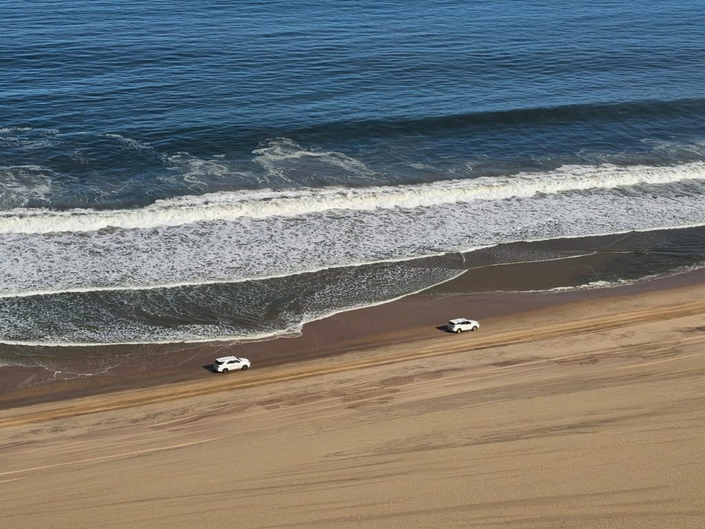 Two white cars drive along the sandy shoreline of a beach. The ocean waves gently crash nearby under a clear blue sky, creating a serene coastal scene.