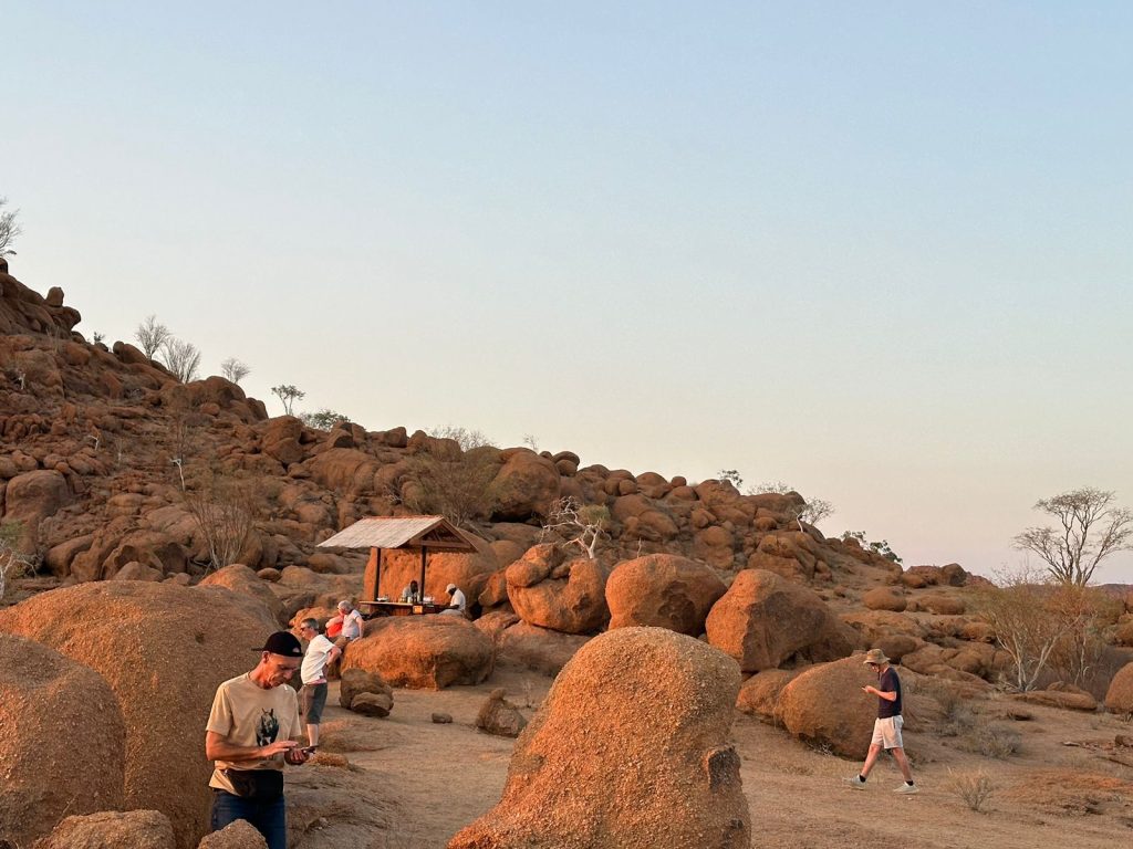 People exploring a rocky landscape at sunset. Some are climbing and taking photos near a small wooden shelter. The scene is dominated by large, warm-hued boulders under a clear sky.