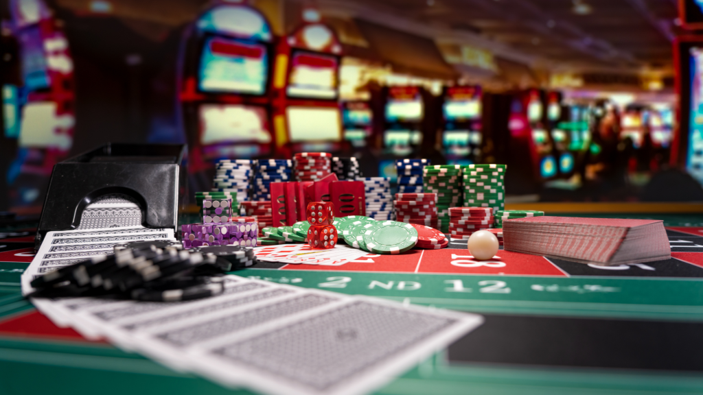 A casino table with playing cards, poker chips, dice, and a deck of cards. Slot machines are blurred in the background, creating a vibrant, lively gambling atmosphere.