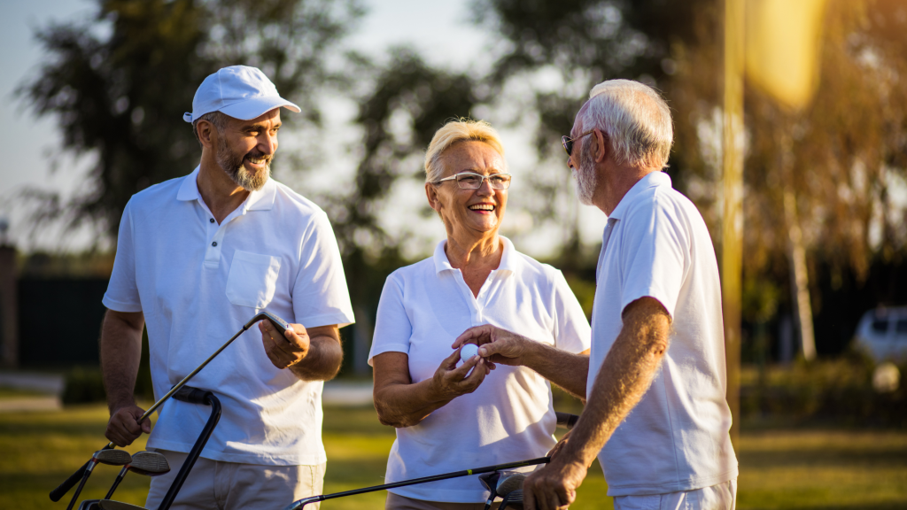 Three older adults, two men and a woman, wearing white shirts and smiling while holding golf clubs. They are standing on a golf course with trees visible in the background. The setting appears relaxed and sunny.