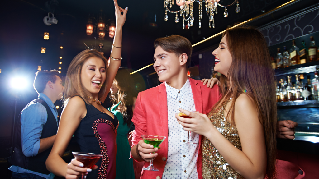 Three friends at a bar, dressed in evening attire, holding cocktails and dancing. They are smiling and appear to be enjoying a lively night out. The background shows shelves with bottles and a warm, festive atmosphere.