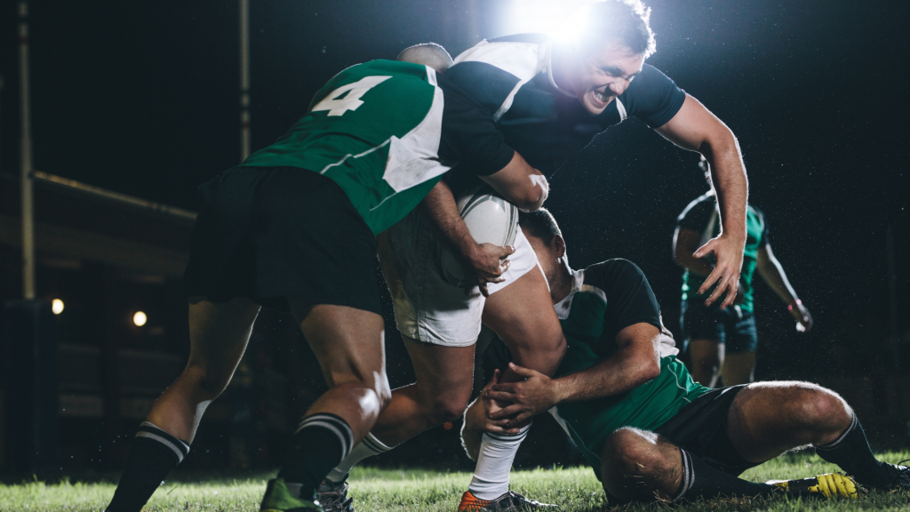 Rugby players in action during a match at night. One player in black and white attempts to break through a tackle, as two opponents in green and white jerseys try to stop him. Stadium lights illuminate the intense scene.