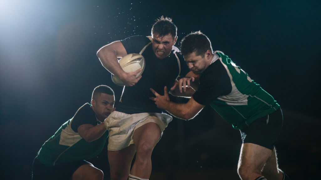 Three rugby players in action during a night game. The central player in black is carrying the ball, fending off two opponents in green jerseys. The intense scene is illuminated by dramatic stadium lighting.