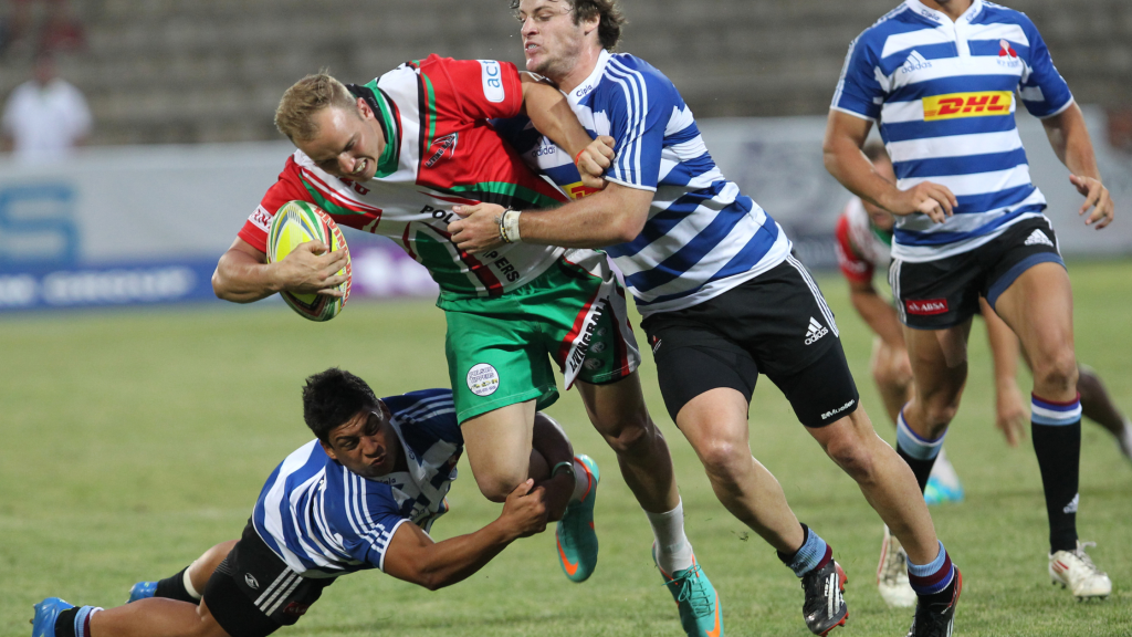 Rugby players in blue and white striped uniforms tackle a player in a red and green uniform who is holding the ball during a match. The scene is dynamic, with players in motion on a grassy field.