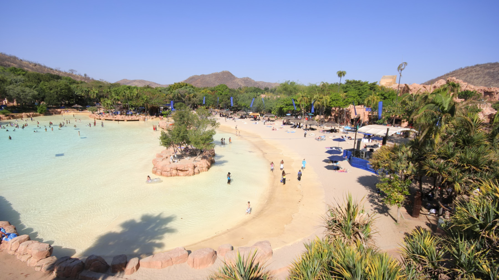 A scenic beach resort featuring a large, clear blue swimming pool surrounded by sand and lush greenery. People are enjoying the water and sunbathing. In the background, hills and trees enhance the natural setting under a clear blue sky.