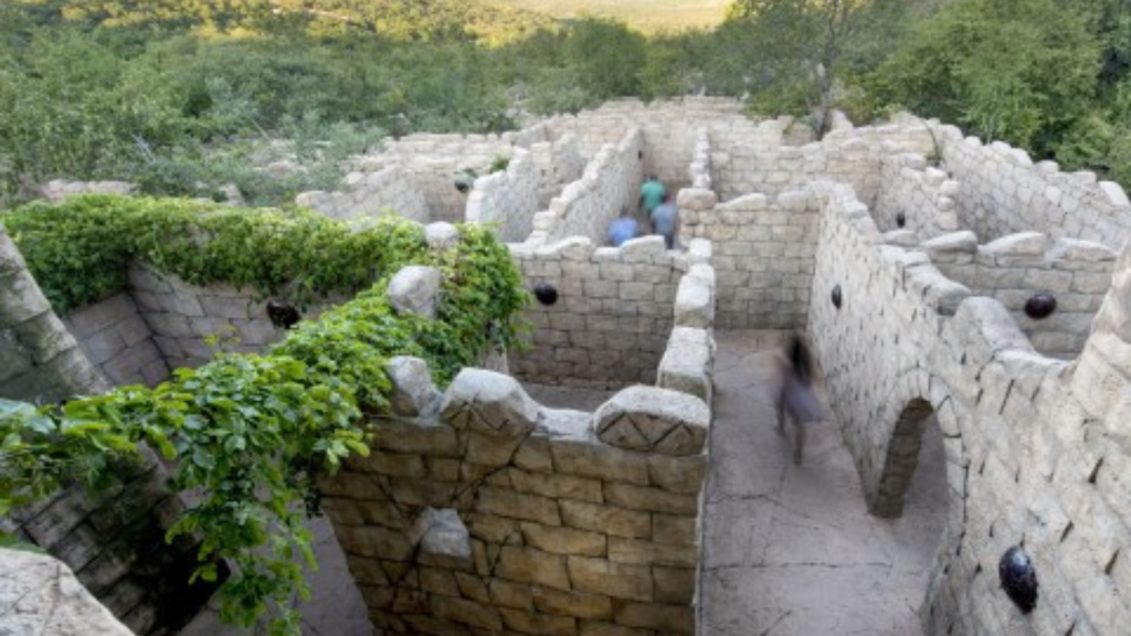 Aerial view of a stone maze with tall walls and green ivy covering parts of the structure. Three people are walking through it, with surrounding lush greenery and hills in the background.