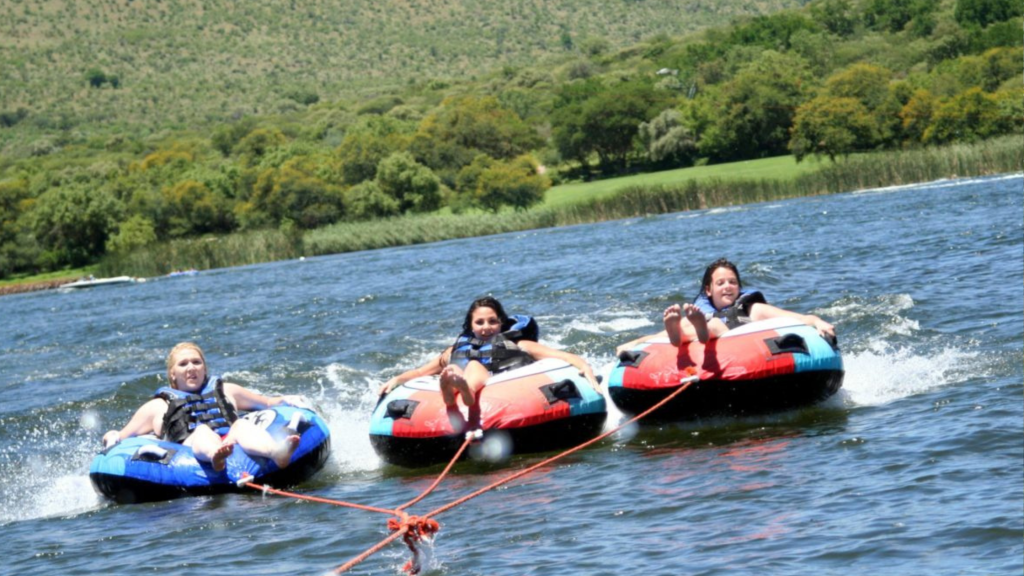 Three people wearing life jackets are joyfully tubing on a lake, each seated on inflatable tubes connected by ropes. The lush green shore and distant hills are visible in the background under a sunny sky.