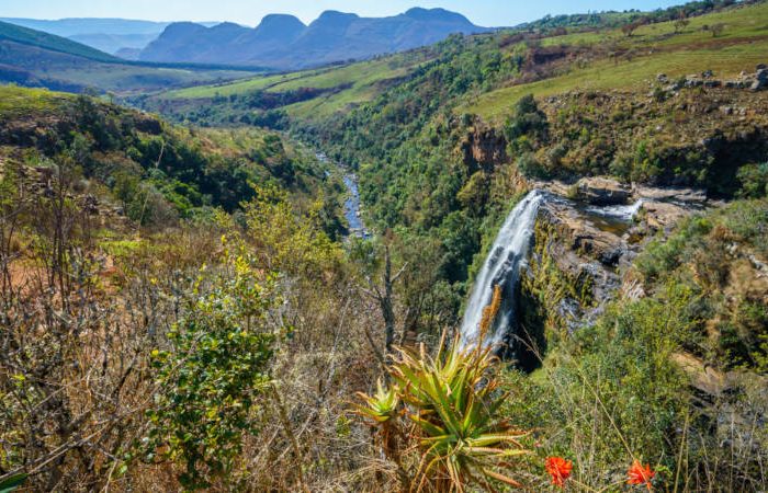 A breathtaking view of the Berlin Falls in South Africa, cascading down a rugged cliff amidst lush greenery. The river winds through a verdant valley, with mountain peaks visible in the distance under a clear blue sky.