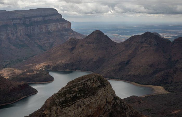 Aerial view of a scenic landscape with rugged mountain ranges and a winding river below. The sky is overcast, casting soft, diffused light over the terrain, highlighting the textures and contours of the rocky formations.