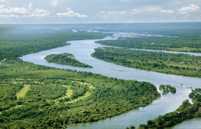 Aerial view of a winding river cutting through a lush green landscape, with dense forests on either side. The sky is partly cloudy, creating a serene and natural scene.