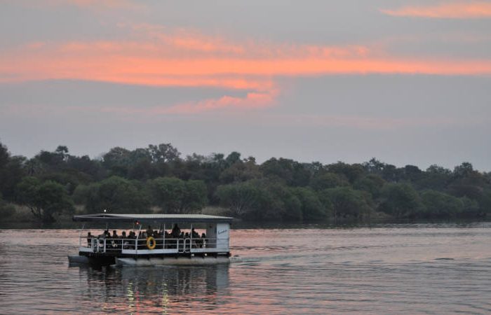 A boat with passengers sails on a calm river during sunset. The sky is painted with shades of orange and pink, and lush green trees line the riverbank, reflecting in the water.