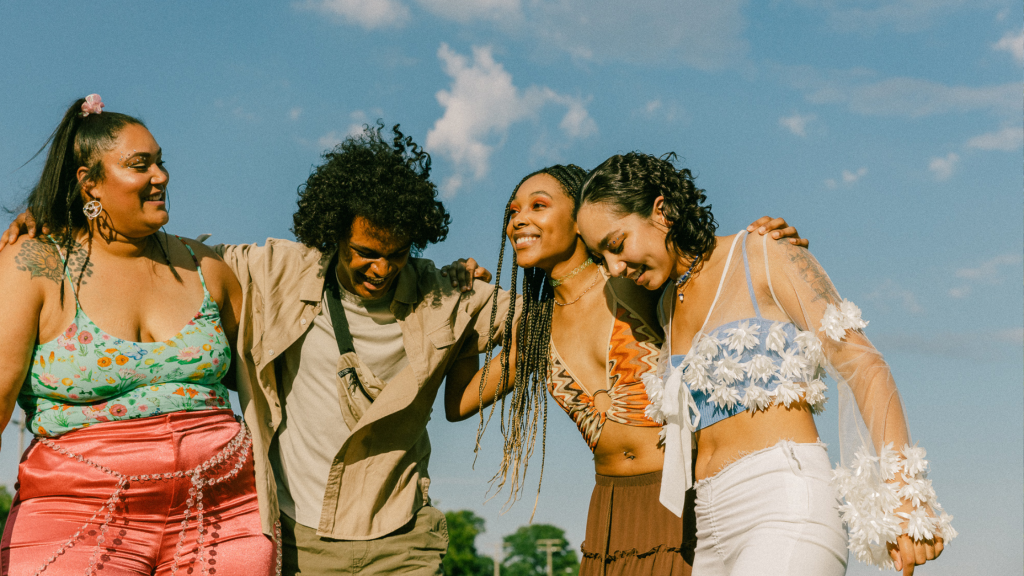 A group of four people stand together outdoors, smiling and embracing, with a bright blue sky and fluffy clouds in the background. They appear happy and relaxed, dressed in colorful summer outfits.