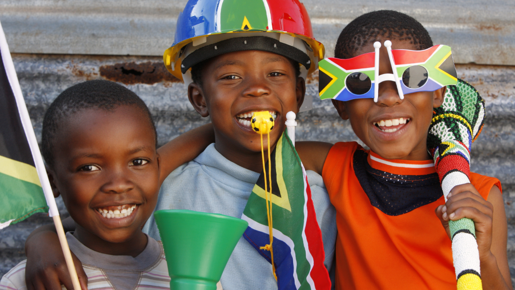 Three smiling children with flags and accessories in South African colors. They wear festive headgear and glasses, and one holds a vuvuzela. The background is a corrugated metal wall, creating a cheerful and lively atmosphere.