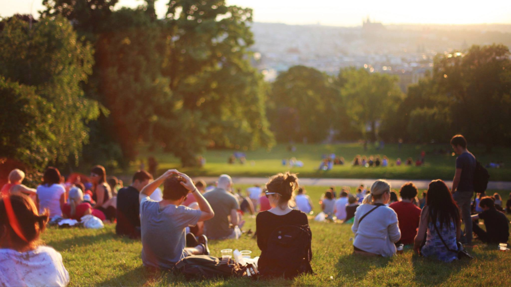 A large group of people sits on a grassy hillside at sunset, enjoying a scenic view. Trees surround the area, and the distant horizon is softly illuminated by the warm light. The atmosphere is relaxed and peaceful.