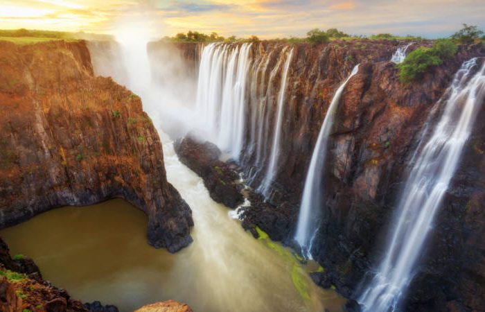 Majestic waterfall cascading over a series of jagged cliffs with lush greenery, creating misty spray and a river below. The scene is vibrant with warm hues, capturing the powerful and serene beauty of nature at sunset.