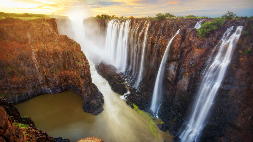 Majestic waterfall cascading over a series of jagged cliffs with lush greenery, creating misty spray and a river below. The scene is vibrant with warm hues, capturing the powerful and serene beauty of nature at sunset.