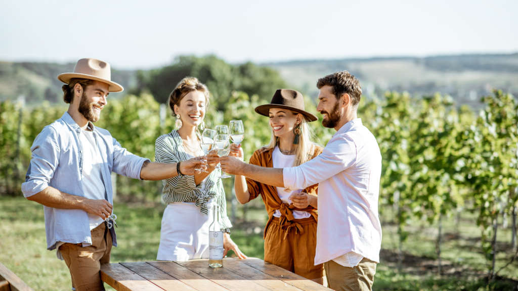 Four people standing outside and clinking glasses of white wine at a wooden table in a vineyard. They are smiling and wearing casual clothing and hats, with a scenic view of grapevines and rolling hills in the background.