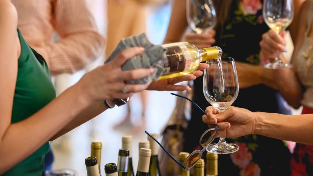 A person pours white wine into a glass held by another person at a social gathering. Several wine bottles are visible on a table, and people are holding wine glasses in the background. The atmosphere suggests a casual or festive occasion.