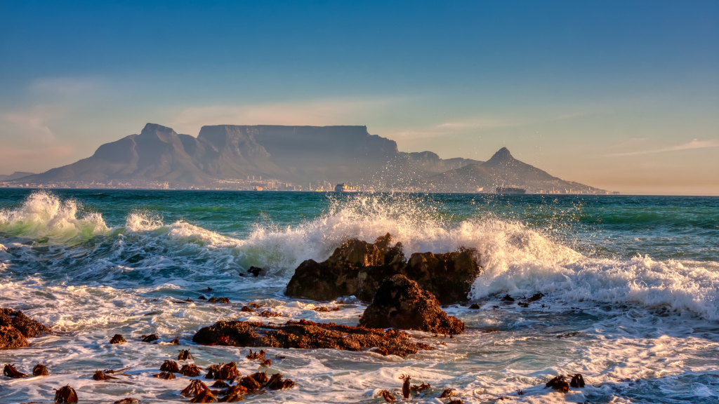 A scenic view of Table Mountain with waves crashing against rocks in the foreground. The mountain is silhouetted against a clear blue sky, and the sea is vibrant with splashes of white foam.