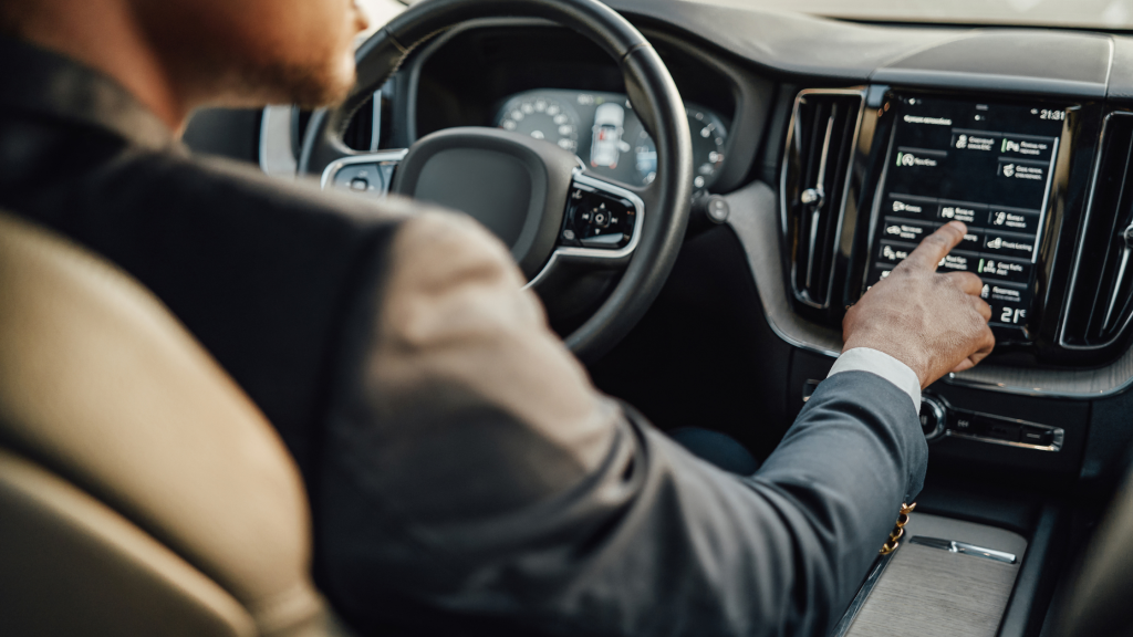 A person in a suit is driving a car and using the touchscreen on the dashboard. The interior is modern, and the driver is focused on the screen which displays various options.