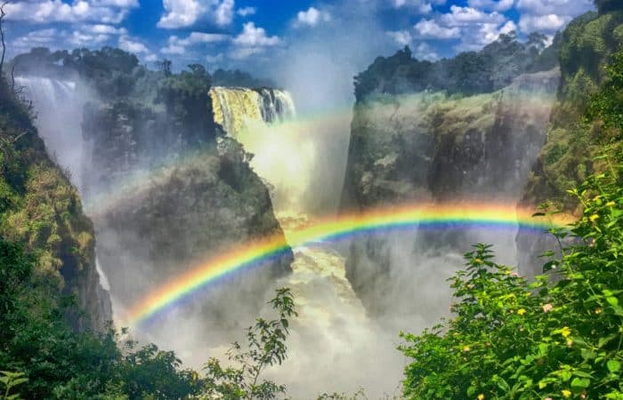 A powerful waterfall surrounded by lush greenery with mist rising into the air. Two vibrant rainbows arch over the chasm, with blue skies and scattered clouds above, creating a vivid and dynamic natural scene.