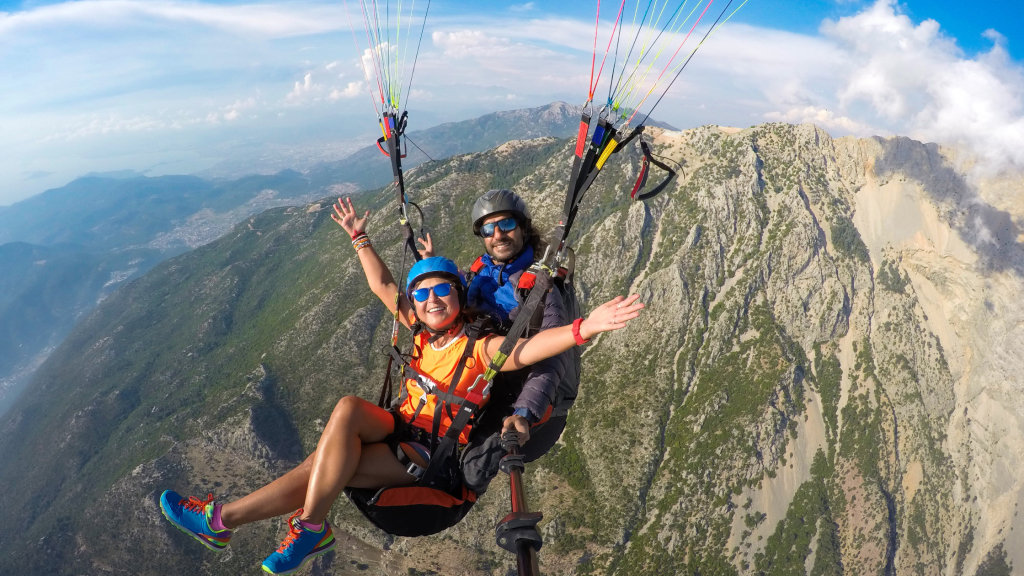 Two people are paragliding over a mountainous landscape with green and rocky terrain. They are smiling, wearing helmets and harnesses, and have their arms raised in excitement. The sky is clear with some clouds in the background.