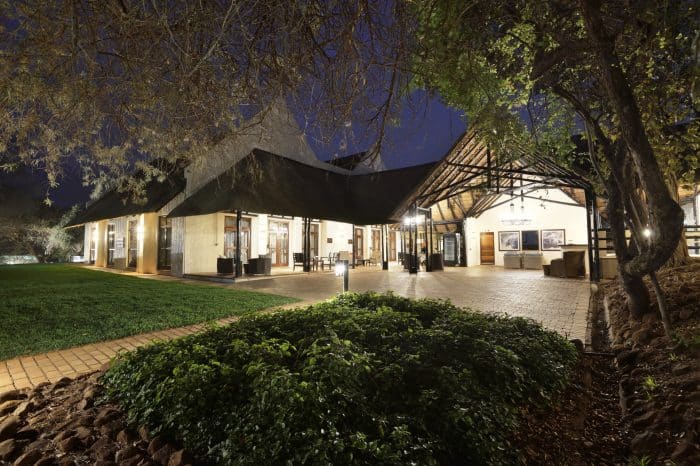 A well-lit, rustic building with a thatched roof is surrounded by trees at night. The entrance is flanked by lit lamps and brick pathways, with greenery in the foreground. The atmosphere is tranquil and inviting.