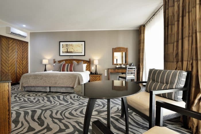 Hotel room with a king-size bed, striped pillows, and a zebra-patterned carpet. Theres a round black table and two chairs in the foreground. A dressing table with a mirror and a framed picture are on the walls, with curtains on the right side.