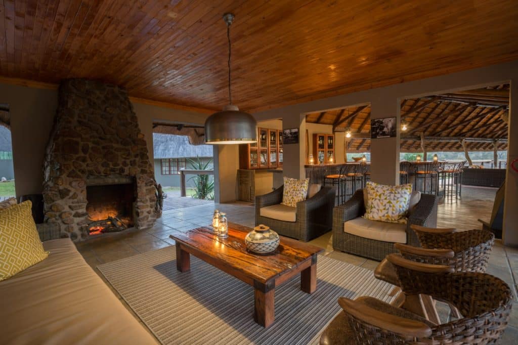 A cozy living room with wooden ceilings and a large stone fireplace. The space features cushioned wicker furniture, a wooden coffee table with candles, and a dining area in the background, creating a warm, inviting atmosphere.