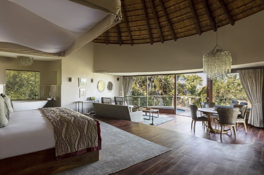 Luxurious open-plan hotel room with a thatched ceiling, featuring a plush bed, lounge area, and dining table. Large windows offer a scenic view of a lush forest. The space is elegantly decorated with neutral tones and a chandelier.